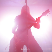 Silhouette of the bass player/vocalist of the band Septicflesh backlit by bright light, glowing white in the center and pink to reddish further out. The silhouette is pink to red due to the lighting.