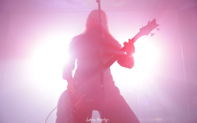 Silhouette of the bass player/vocalist of the band Septicflesh backlit by bright light, glowing white in the center and pink to reddish further out. The silhouette is pink to red due to the lighting.