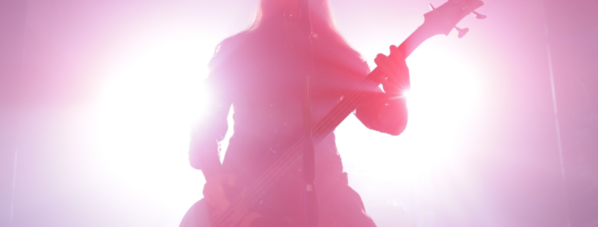 Silhouette of the bass player/vocalist of the band Septicflesh backlit by bright light, glowing white in the center and pink to reddish further out. The silhouette is pink to red due to the lighting.