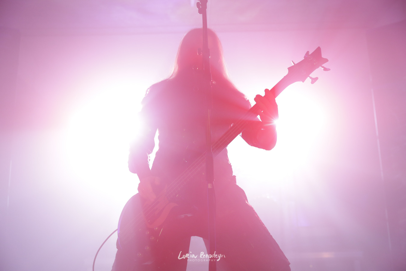 Silhouette of the bass player/vocalist of the band Septicflesh backlit by bright light, glowing white in the center and pink to reddish further out. The silhouette is pink to red due to the lighting.